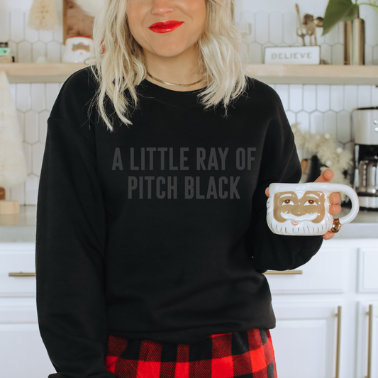 A person with blonde hair, in a unisex Pitch Black • Tonal Pullover reading A LITTLE RAY OF PITCH BLACK and a red plaid skirt, holds a Santa face mug in a white kitchen.