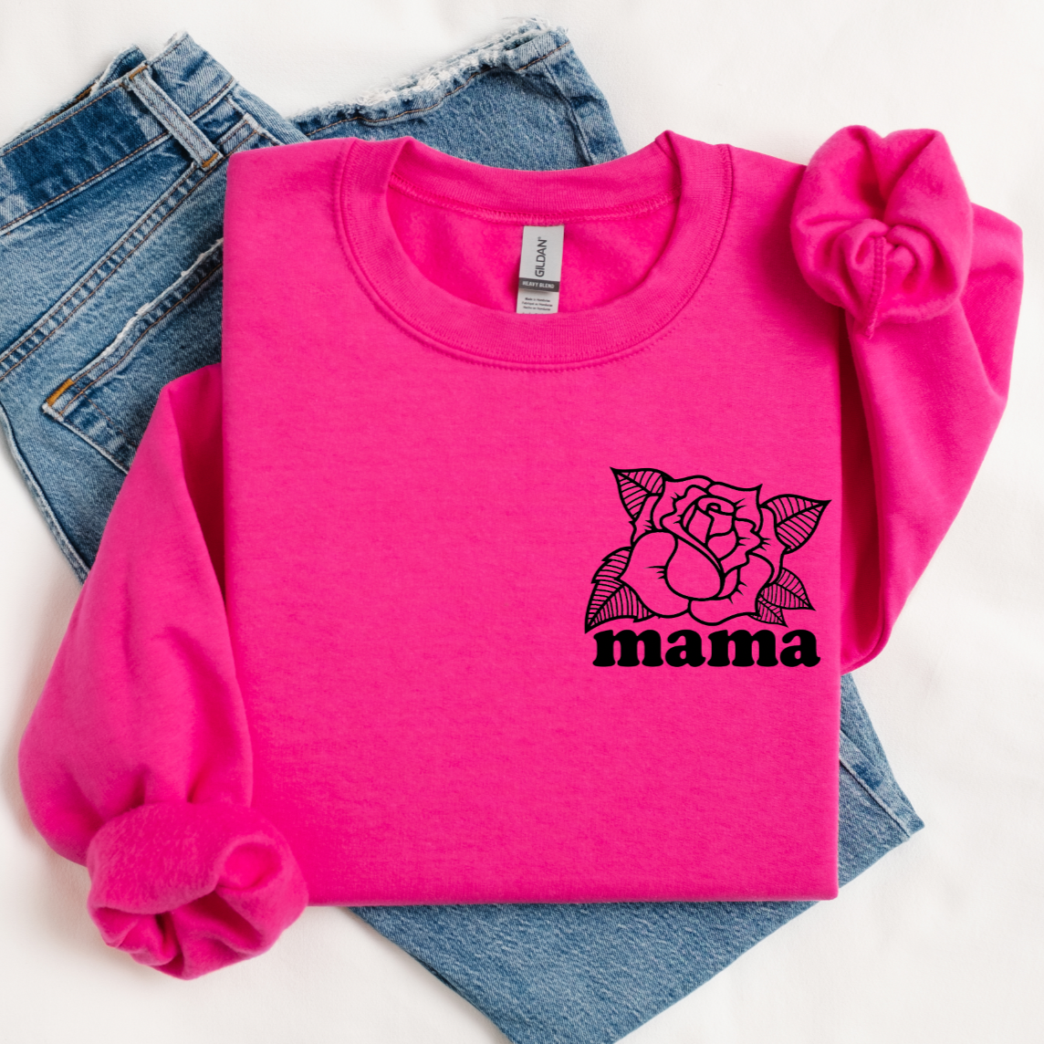 The Mama Rose • Cyber Pink Pullover, featuring a black rose graphic on the chest, is folded atop light blue jeans and paired with a pink scrunchie—perfect for Valentine’s Day in a comfy unisex fit.