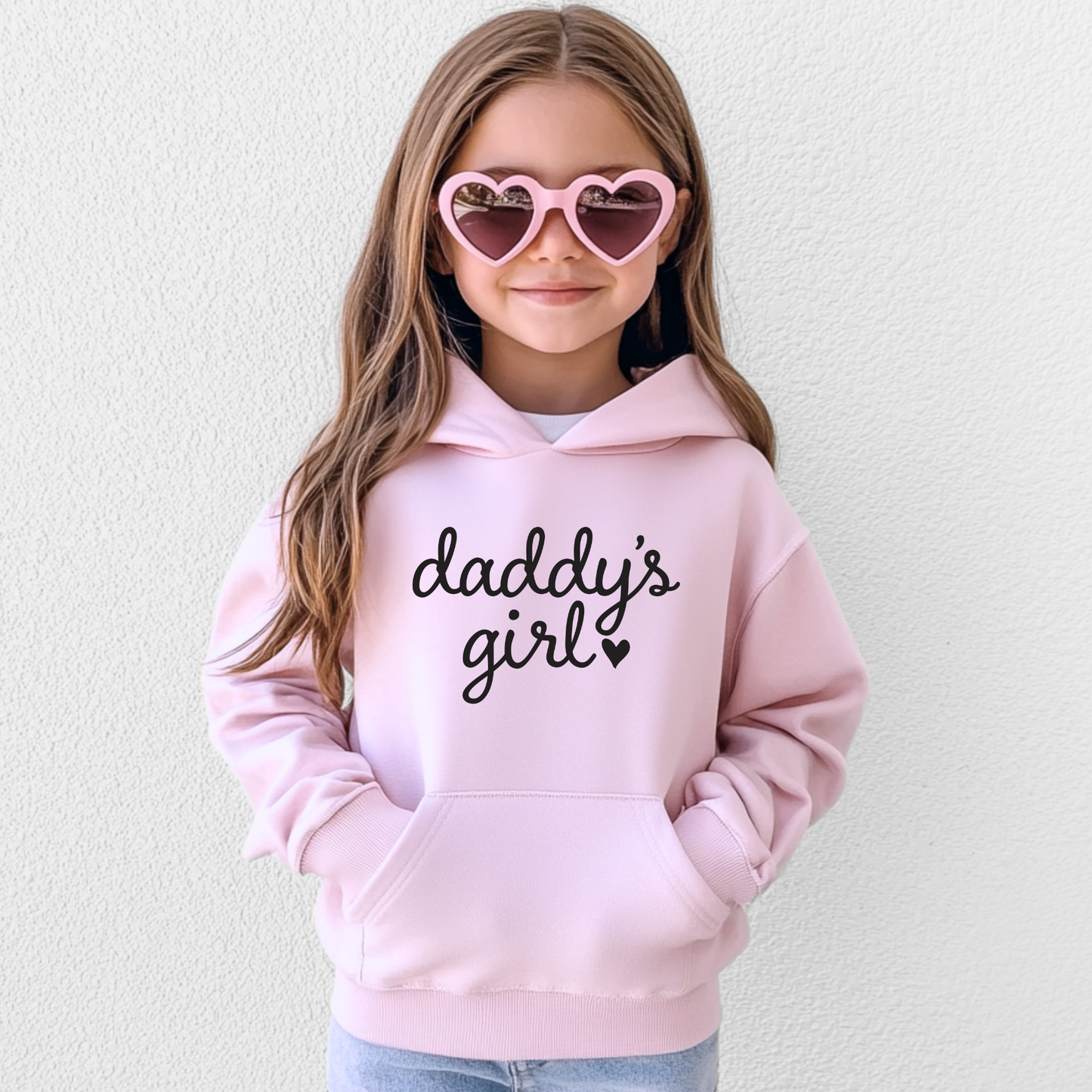 A young girl with long brown hair wears heart-shaped pink sunglasses and the Daddys Girl • Pink Hoodie. She stands against a plain white background, smiling slightly.