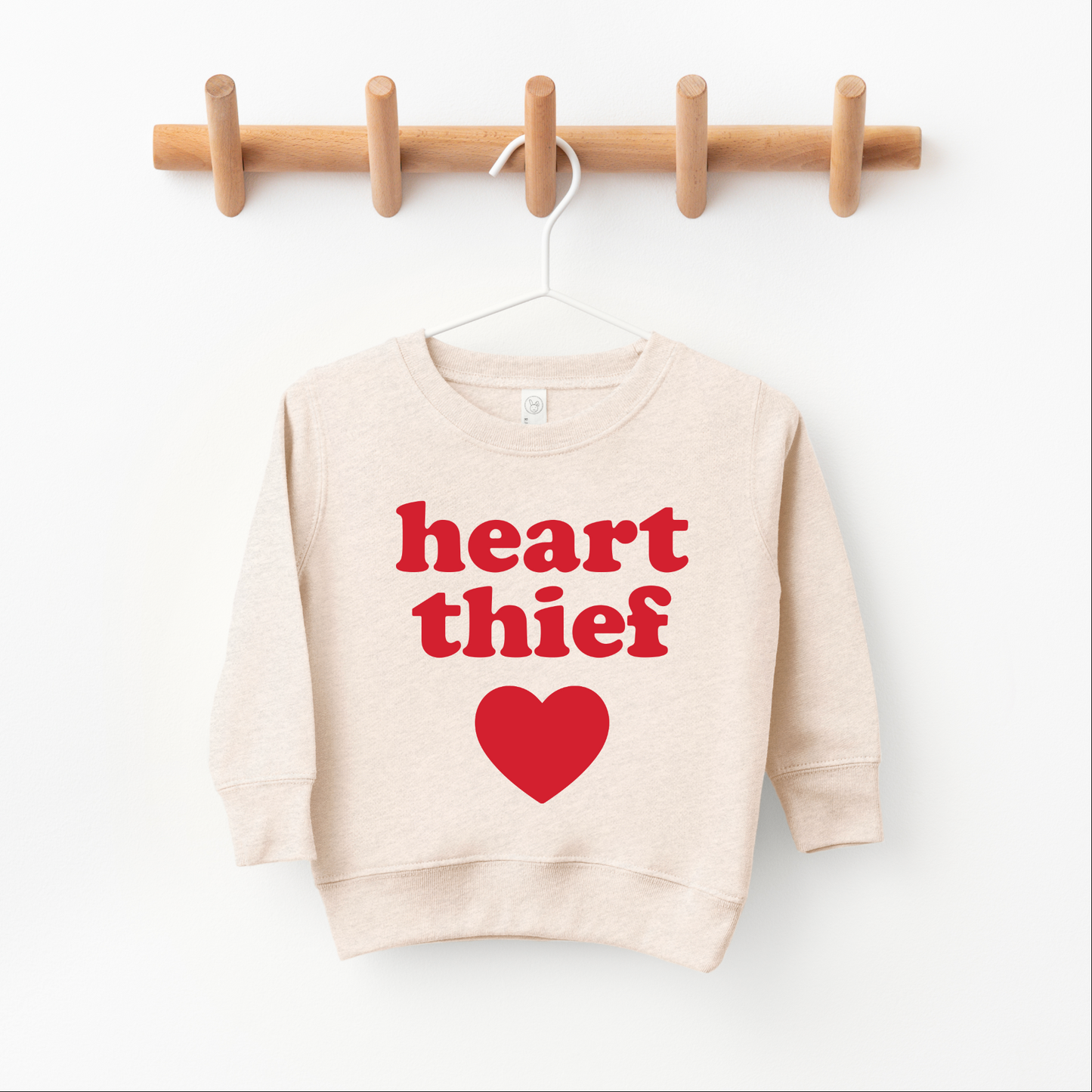 The Heart Thief • Oatmeal Pullover is a unisex kids’ sweatshirt with bold red “heart thief” text and a large heart graphic, hanging on a wooden peg rack. For comfort as your child grows, we recommend sizing up.