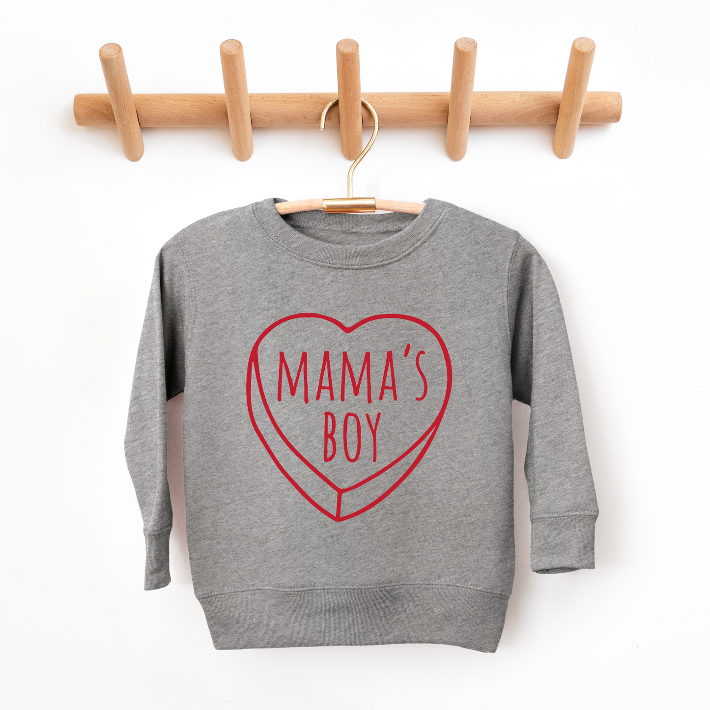 The Mamas Boy • Graphite Pullover is a gray unisex kid’s sweatshirt with a red heart outline and “MAMAS BOY” text, shown on a hanger and rack against a white wall. Please check size-up instructions for the best fit.