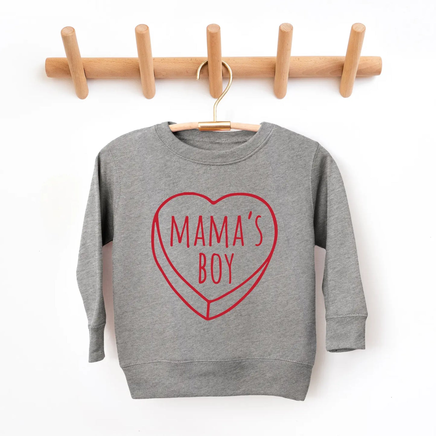 The Mamas Boy • Graphite Pullover is a gray unisex kid’s sweatshirt with a red heart outline and “MAMAS BOY” text, shown on a hanger and rack against a white wall. Please check size-up instructions for the best fit.