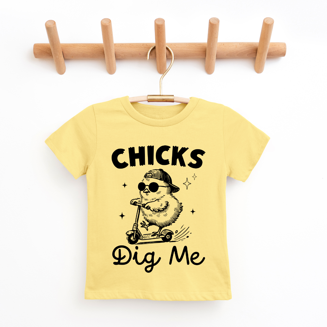 A yellow Chicks Dig Me • Yellow Tee for kids hangs on a wooden rack, featuring a cartoon chick in sunglasses and cap riding a scooter, with “Chicks Dig Me” text—crafted from super-soft fabric, perfect for Easter.
