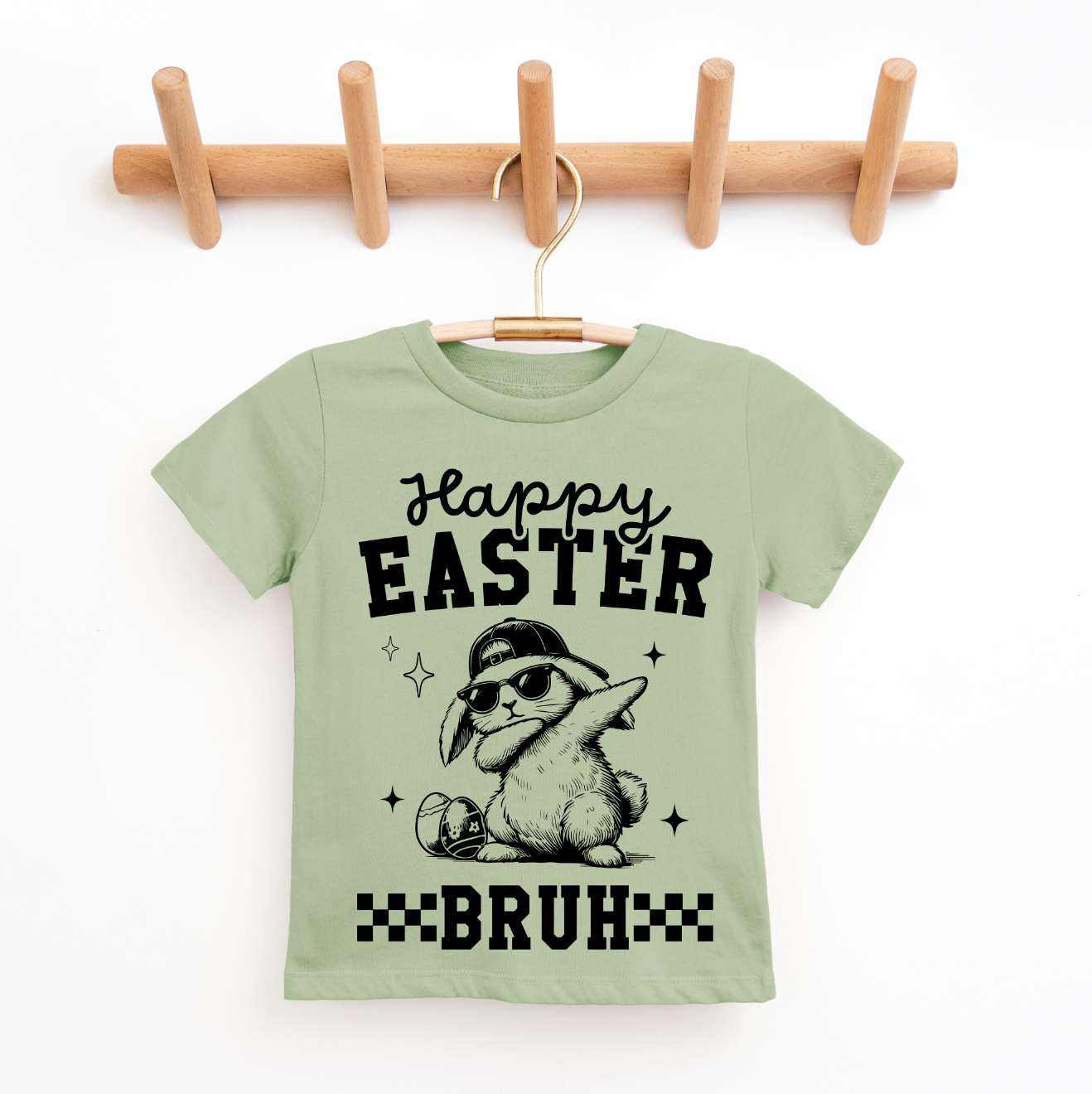 A Happy Easter Bruh • Sage Tee for kids in light green hangs on a wooden rack, showcasing a cool bunny with sunglasses and cap, “Happy Easter BRUH” text, stars, checkerboard patterns, and fun super-soft fabric.