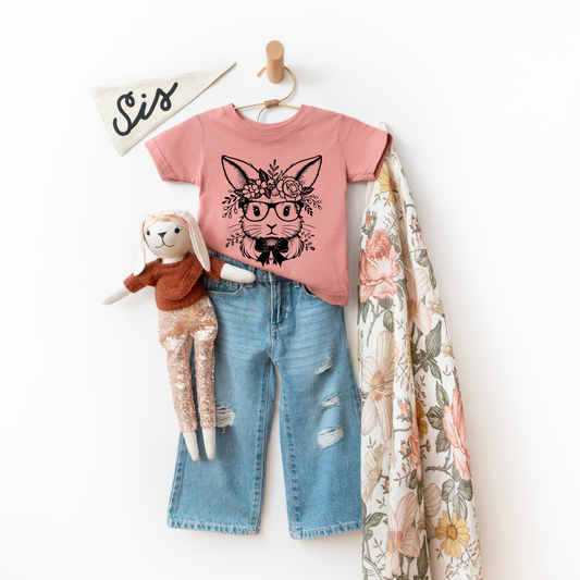 The Floral Bunny • Mauve Tee is a children’s super-soft, 100% combed ring-spun cotton T-shirt in pink featuring a glasses-wearing rabbit, paired with ripped blue jeans, a plush bunny doll, floral blanket, and “Sis” pennant decor.