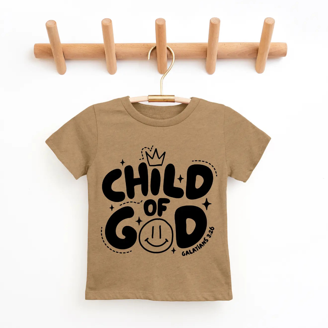 The Child of God • Coyote Tee, a tan unisex childrens shirt, hangs on a wooden rack. It displays bold black CHILD OF GOD text with a crown, stars, smiley face, and Galatians 3:26—crafted with socially conscious care.