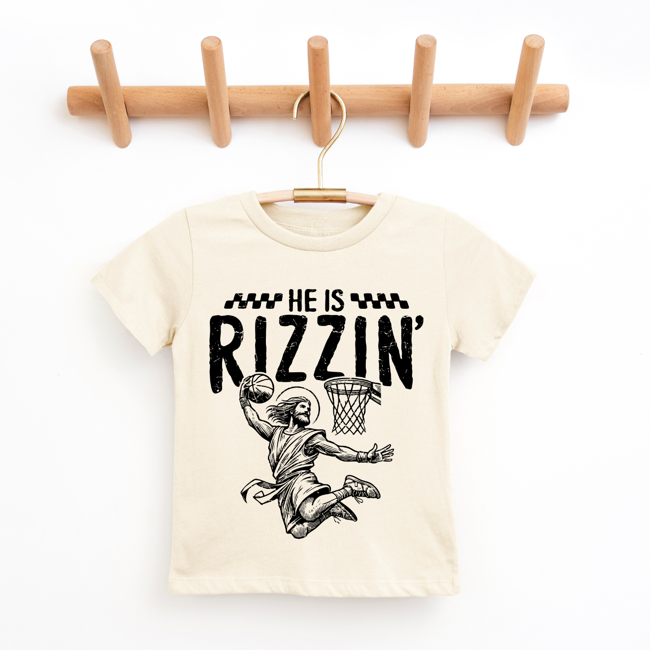 The He is Rizzin • Kids Natural Tee, in beige and hung on a wooden hanger, shows a basketball player dunking with “He is Rizzin’” above—crafted from super-soft fabric for Easter or everyday wear.