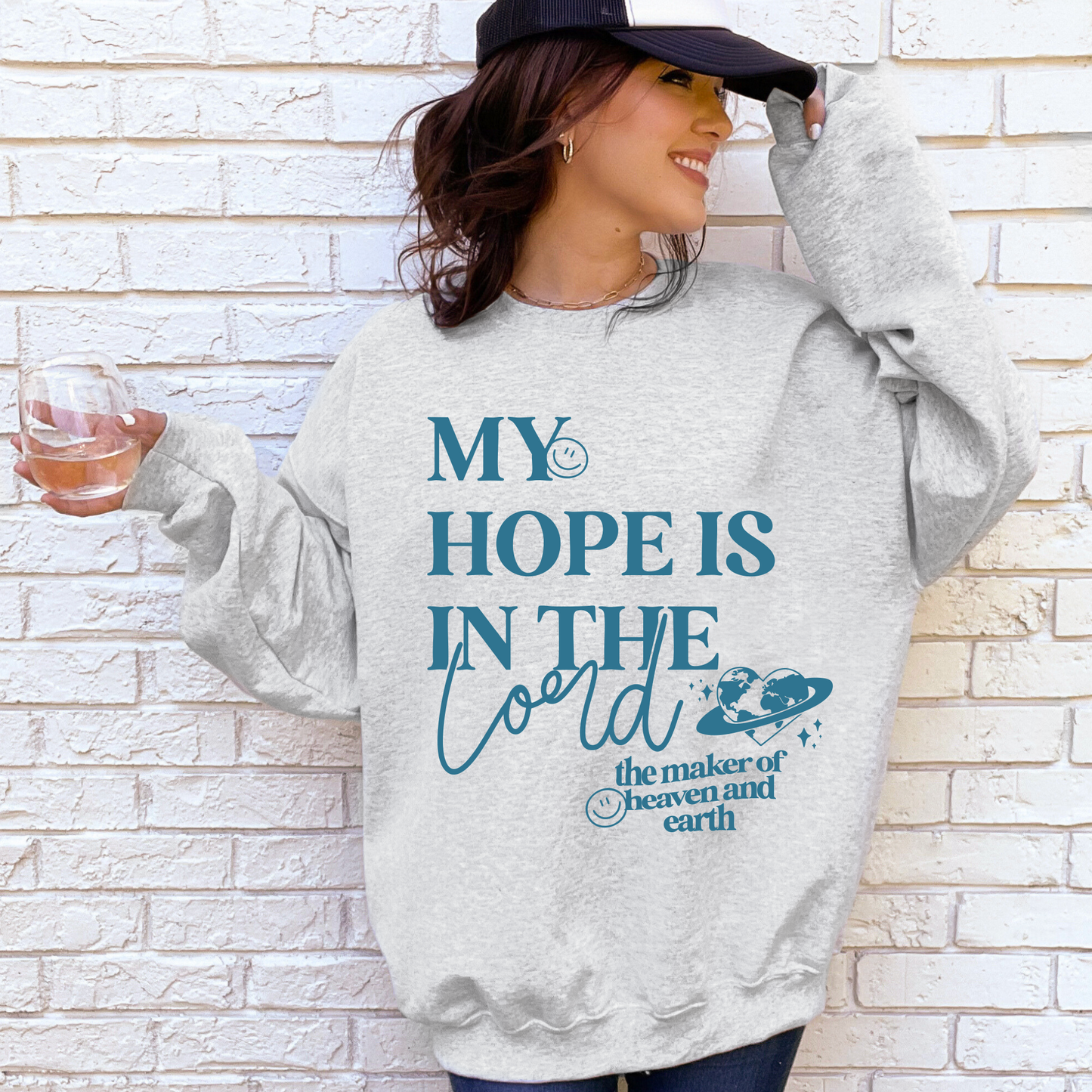 A woman in an ash My Hope • Ash Tee or Pullover smiles, holding a glass of water. Her unisex sweatshirt features the phrase My hope is in the Lord with decorative text and planet illustrations.