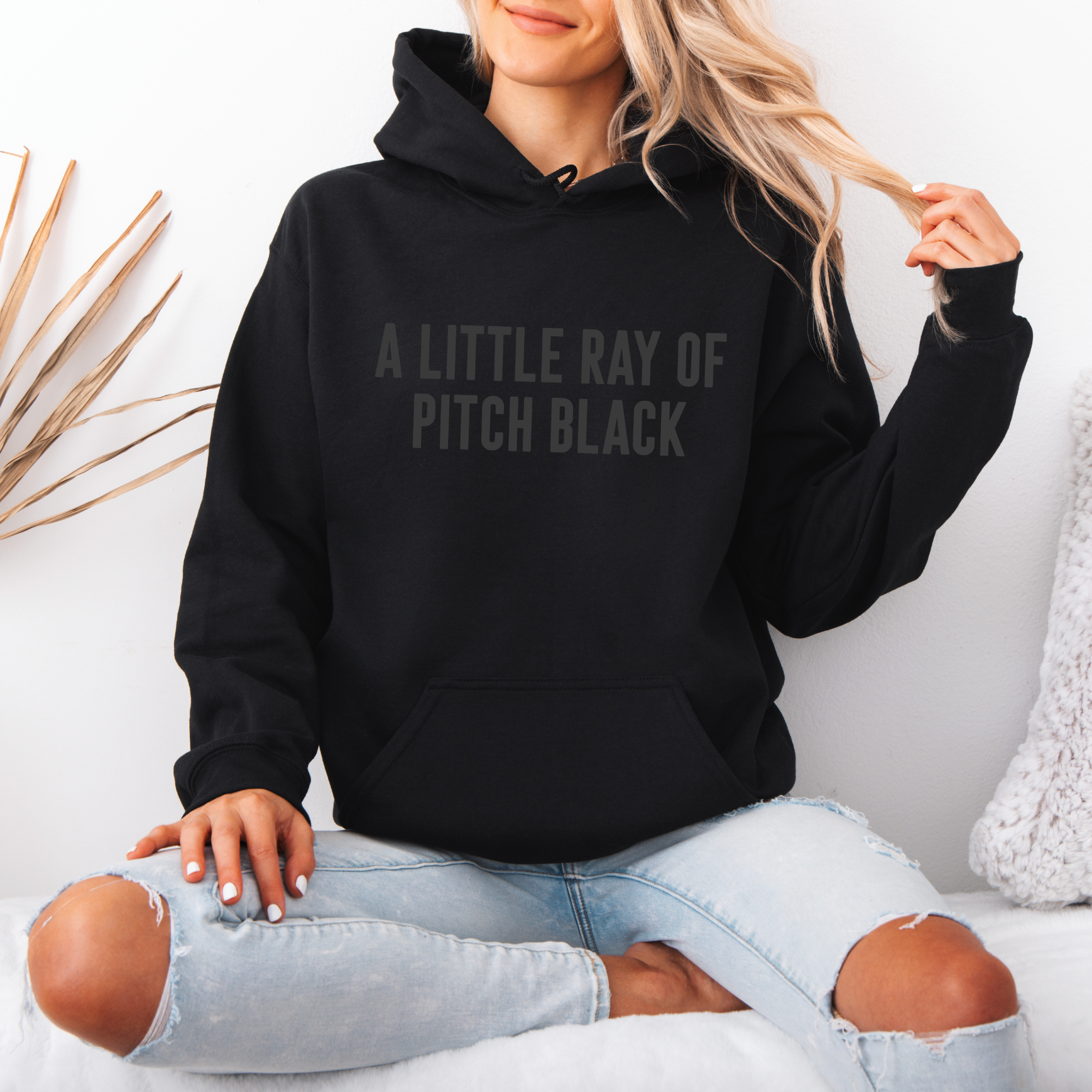A woman with long blonde hair sits cross-legged on a white bed, wearing the Ray of Pitch Black • Tonal Hoodie featuring A LITTLE RAY OF PITCH BLACK text. She pairs this comfy unisex fit with ripped light blue jeans.