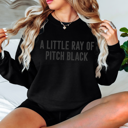A woman with long, wavy blonde hair wears the Pitch Black • Tonal Pullover, featuring the phrase A LITTLE RAY OF PITCH BLACK in subtle dark letters. She sits on a bed, displaying rings and a star necklace.