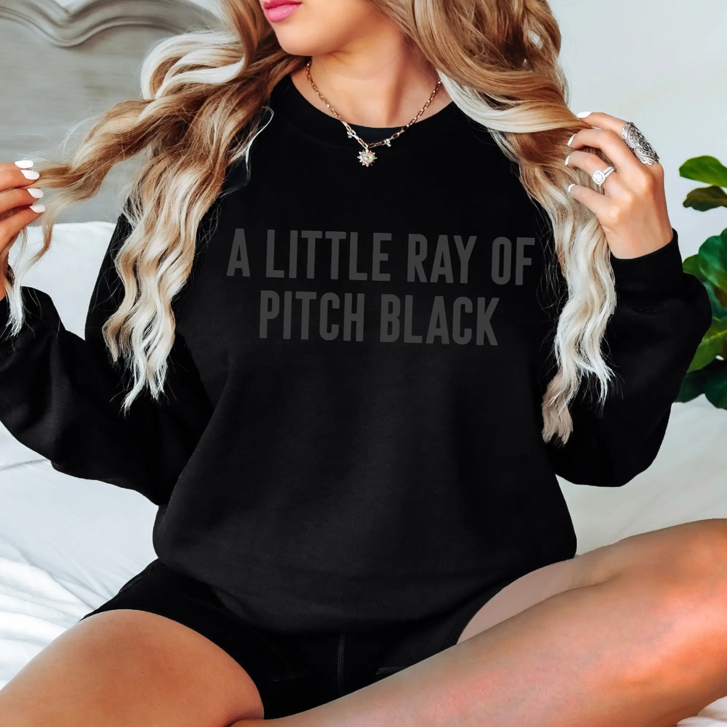 A woman with long, wavy blonde hair wears the Pitch Black • Tonal Pullover, featuring the phrase A LITTLE RAY OF PITCH BLACK in subtle dark letters. She sits on a bed, displaying rings and a star necklace.
