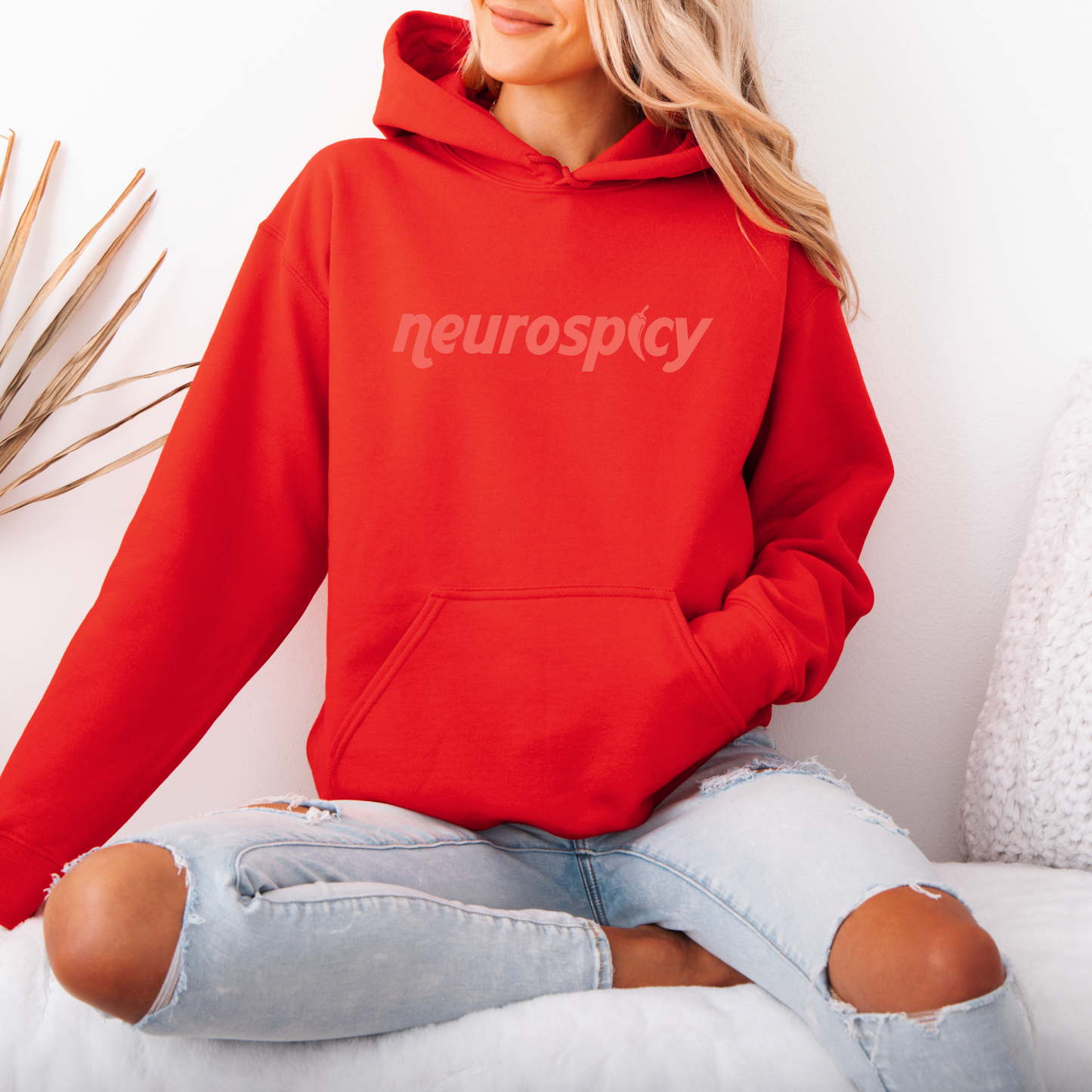 A person with long blonde hair sits cross-legged on a white surface, wearing a bright red neurospicy • Tee or Hoodie and light ripped jeans. A decorative plant is in the background.