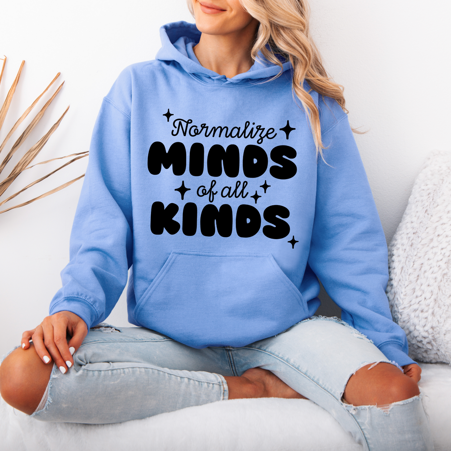 A woman with long blonde hair sits cross-legged in a cozy, minimal space, wearing a light blue Minds of all Kinds • Tee or Hoodie with bold black lettering. She appears relaxed and comfortable.