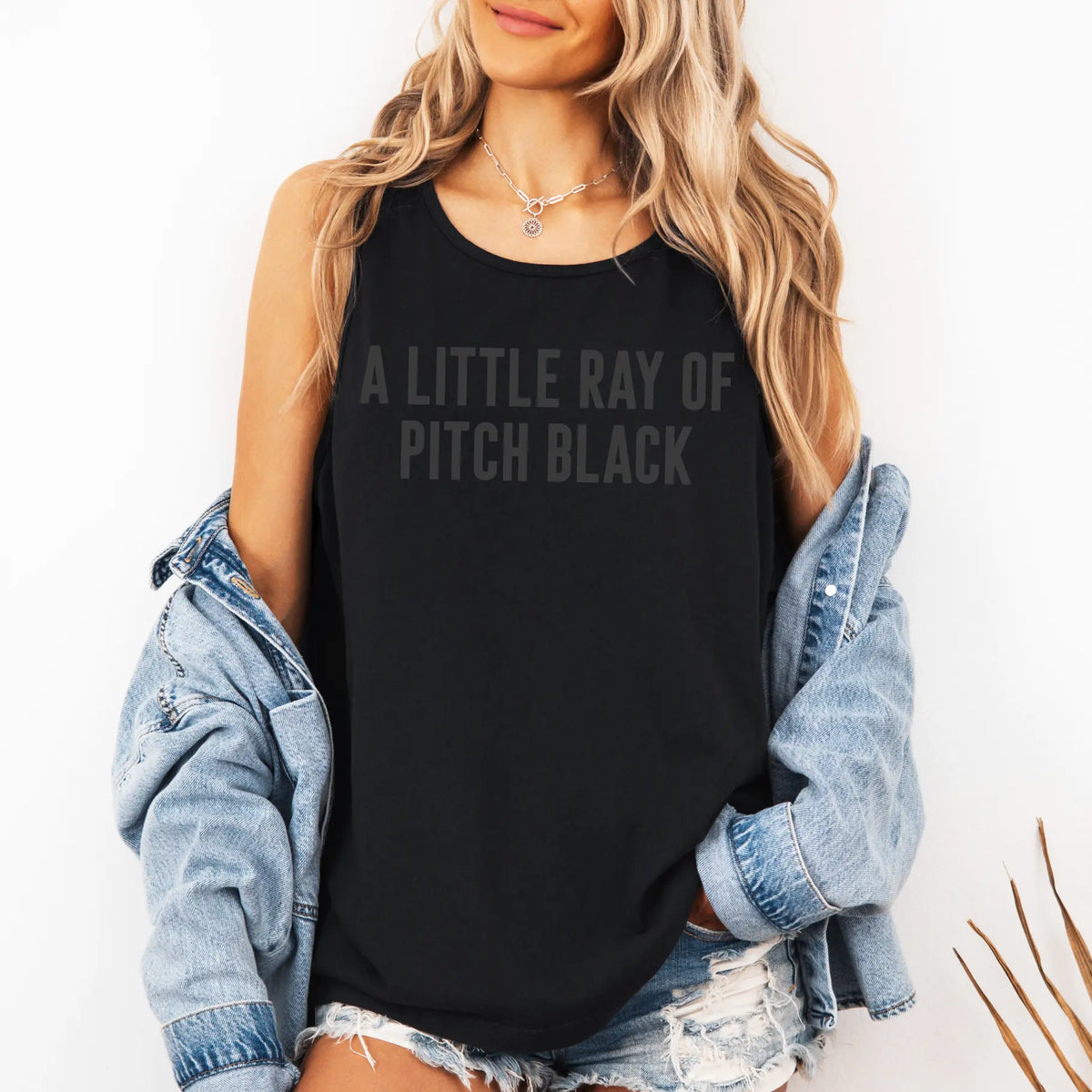 A woman with long blonde hair models the Pitch Black • Unisex Tank Top featuring A LITTLE RAY OF PITCH BLACK text, paired with a denim jacket and shorts, standing against a white background.
