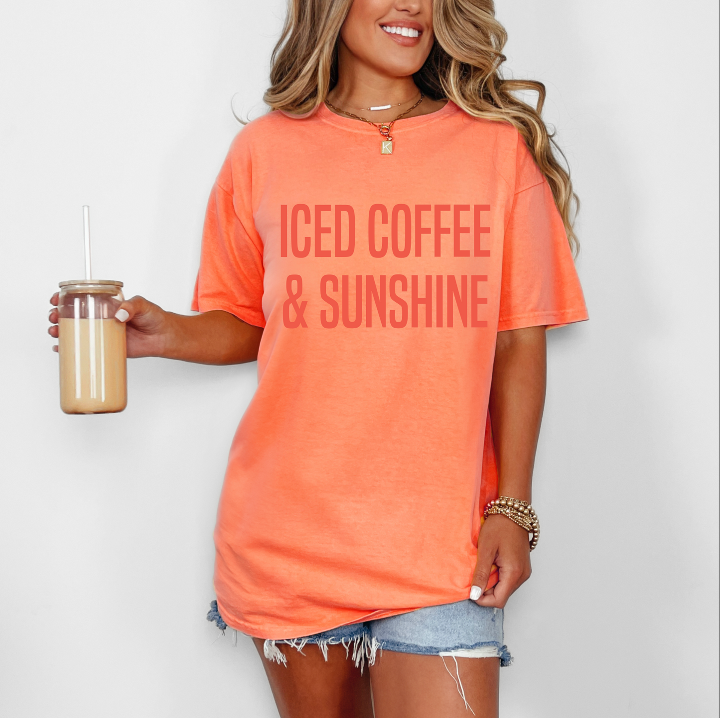 A woman wears the Iced Coffee • Comfort Colors Tee in bright orange, featuring ICED COFFEE & SUNSHINE, made from soft ring-spun cotton, paired with denim shorts and gold accessories while holding an iced coffee against a white background.