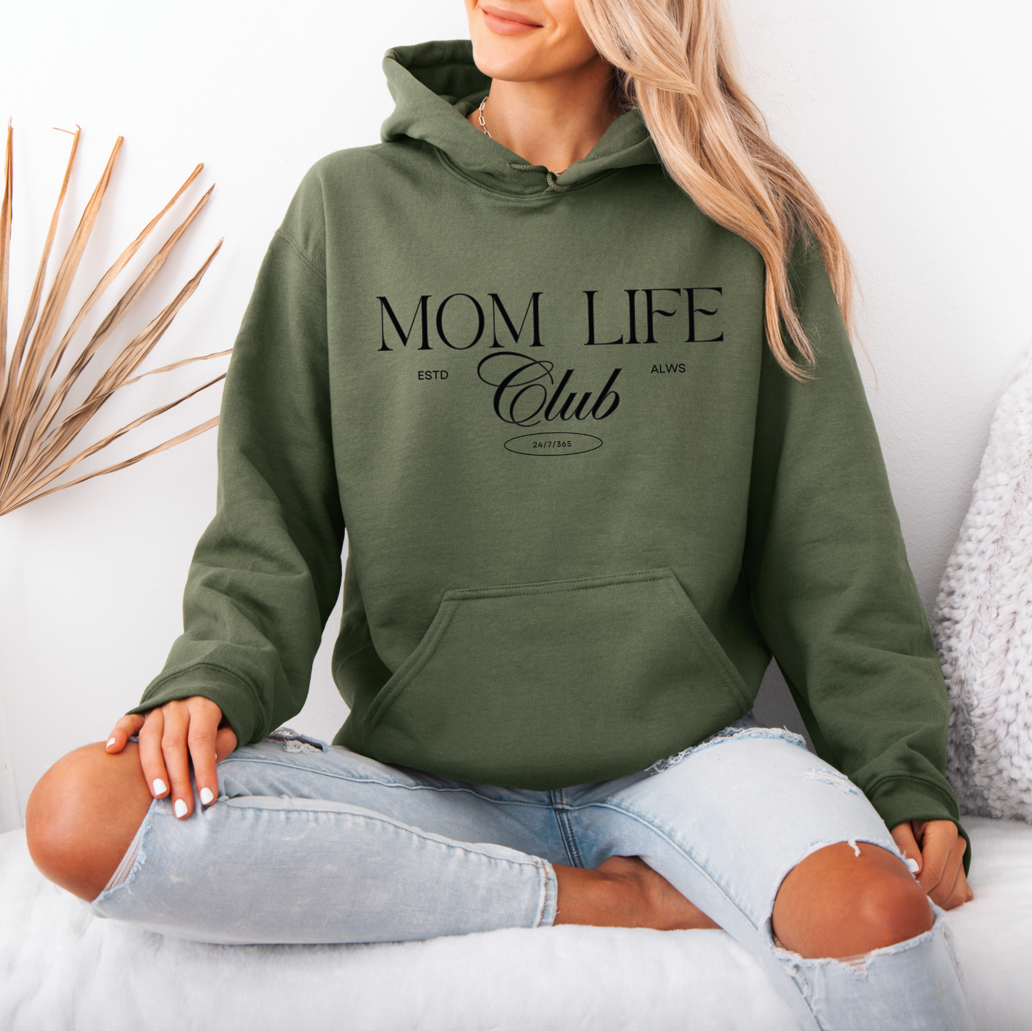 A woman sits cross-legged on a white surface, wearing the Mom Life Club • Military Green Hoodie with light blue ripped jeans. Dried grass decor and a white wall are in the background.