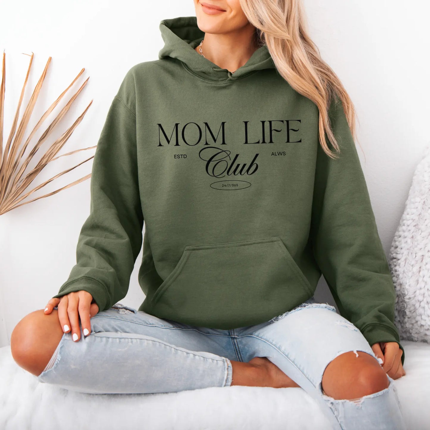 A woman sits cross-legged on a white surface, wearing the Mom Life Club • Military Green Hoodie with light blue ripped jeans. Dried grass decor and a white wall are in the background.