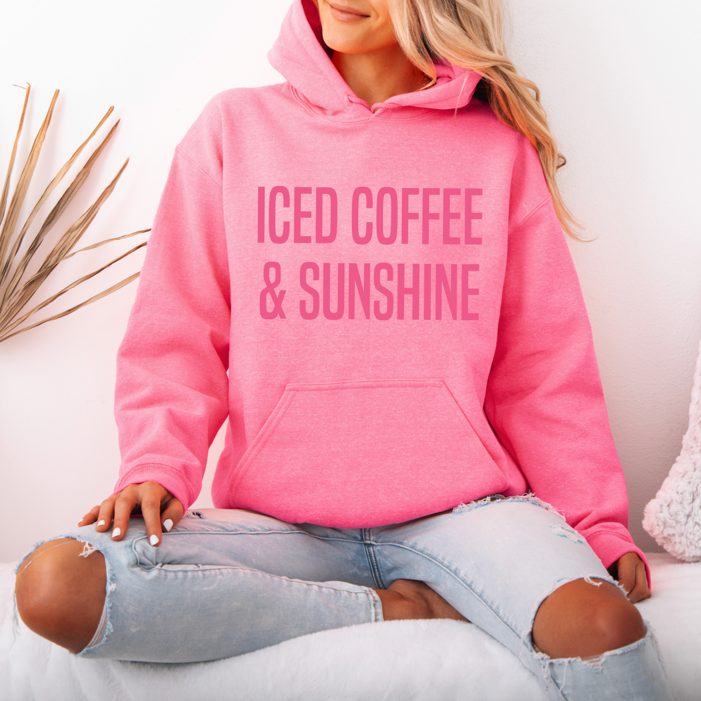 A woman with long blonde hair, dressed in ripped light blue jeans and the Iced Coffee & Sunshine • Neon Pink Hoodie, sits cross-legged on a white couch.