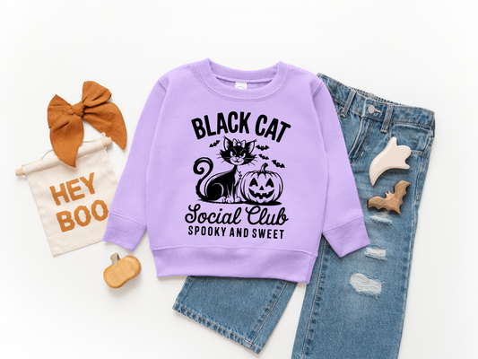 Flat lay of the Black Cat Social Club • Kids Pullover in lavender with a black cat and pumpkin graphic, reading Spooky and Sweet. Styled with ripped jeans, a brown hair bow, toy pumpkins, bats, a Hey Boo sign; available in unisex sizing.