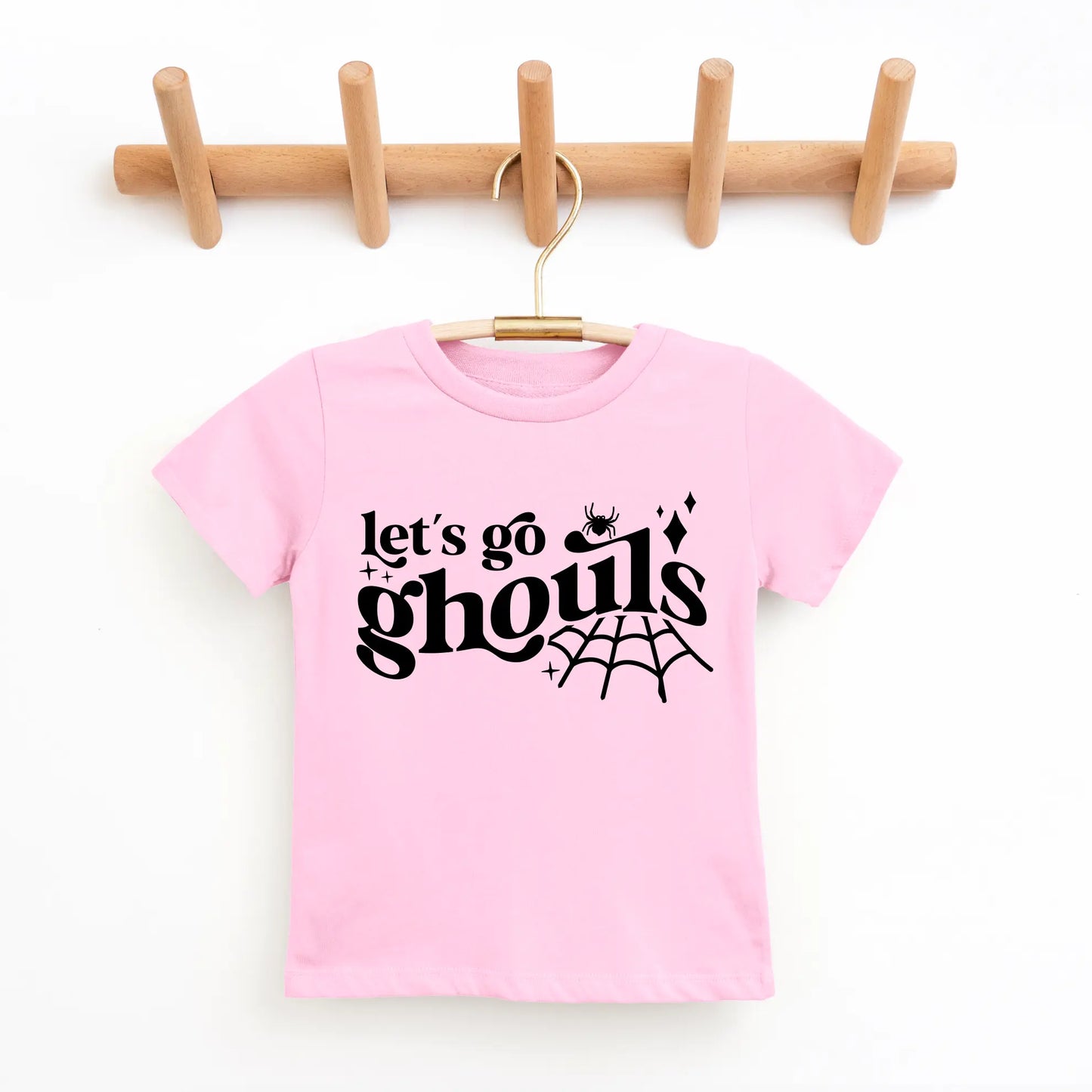 A pink Lets Go Ghouls • Kids Tee made from super-soft cotton hangs on a wooden hook rack, with black text, a small spider, sparkles, and a spiderweb—perfect for kids who love fun and spooky shirts.