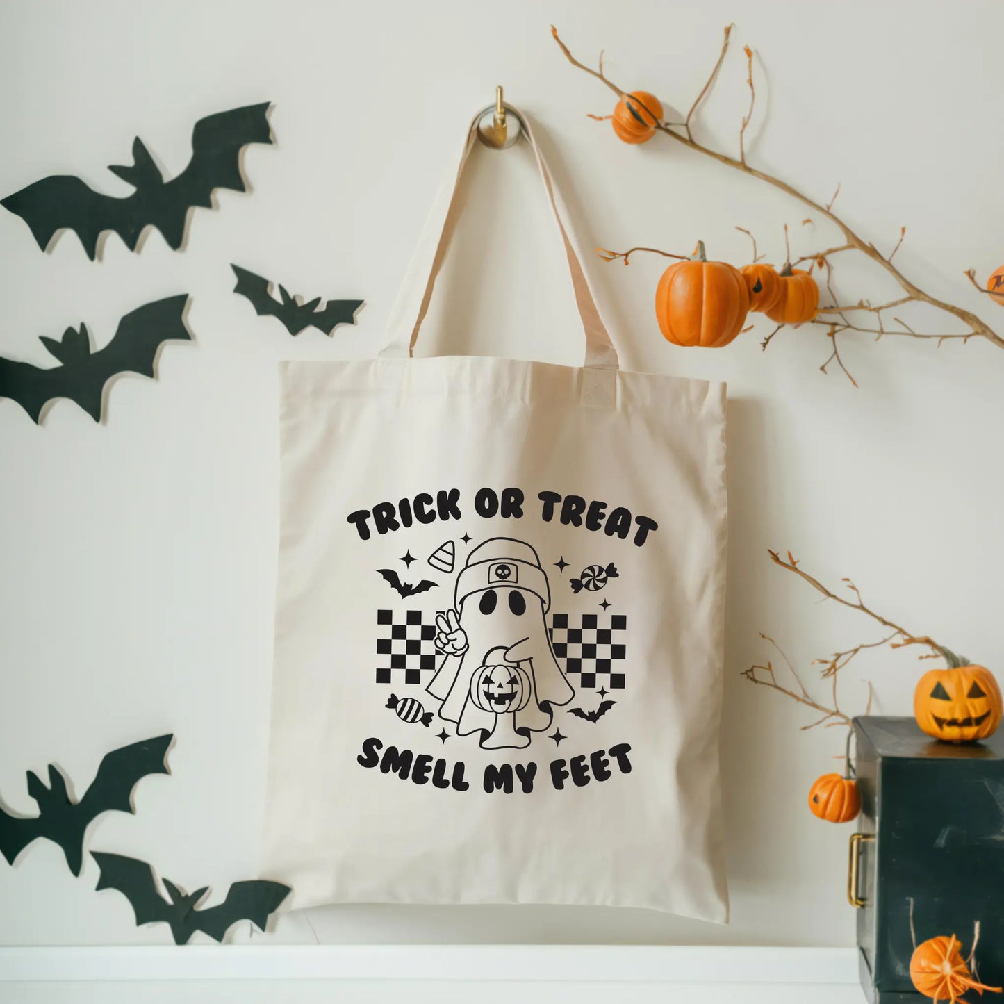 The Trick or Treat Tote, made from sustainable 100% cotton canvas, hangs on a wall with a cartoon ghost, candy, and Trick or Treat Smell My Feet text. Black paper bats and pumpkin decorations complete the Halloween look.