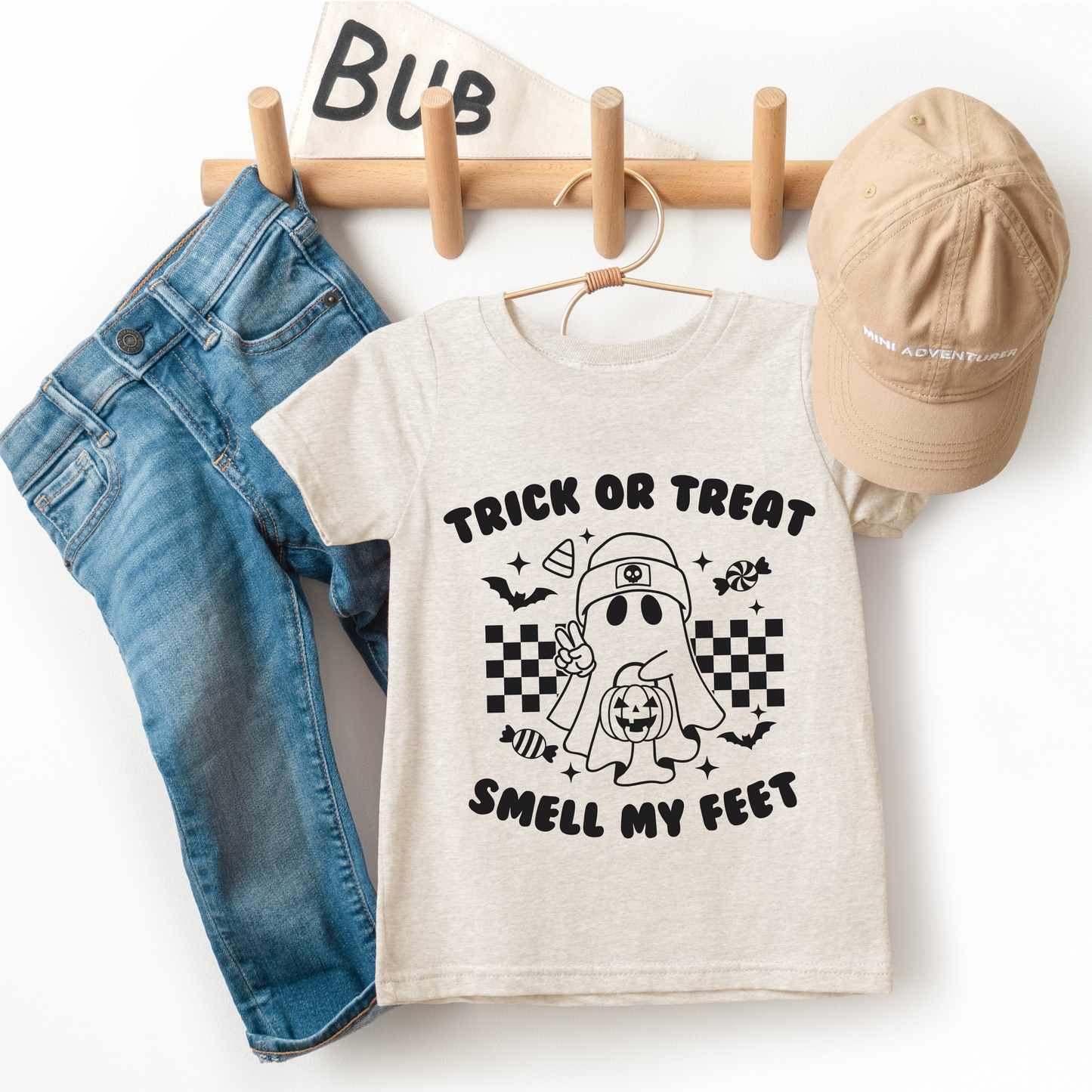 The Smell My Feet • Kids Tee is a beige unisex t-shirt featuring a cartoon ghost and Trick or Treat Smell My Feet text, made from super-soft cotton, shown with blue jeans and a beige cap on a wooden rack beside a “Bub” flag.