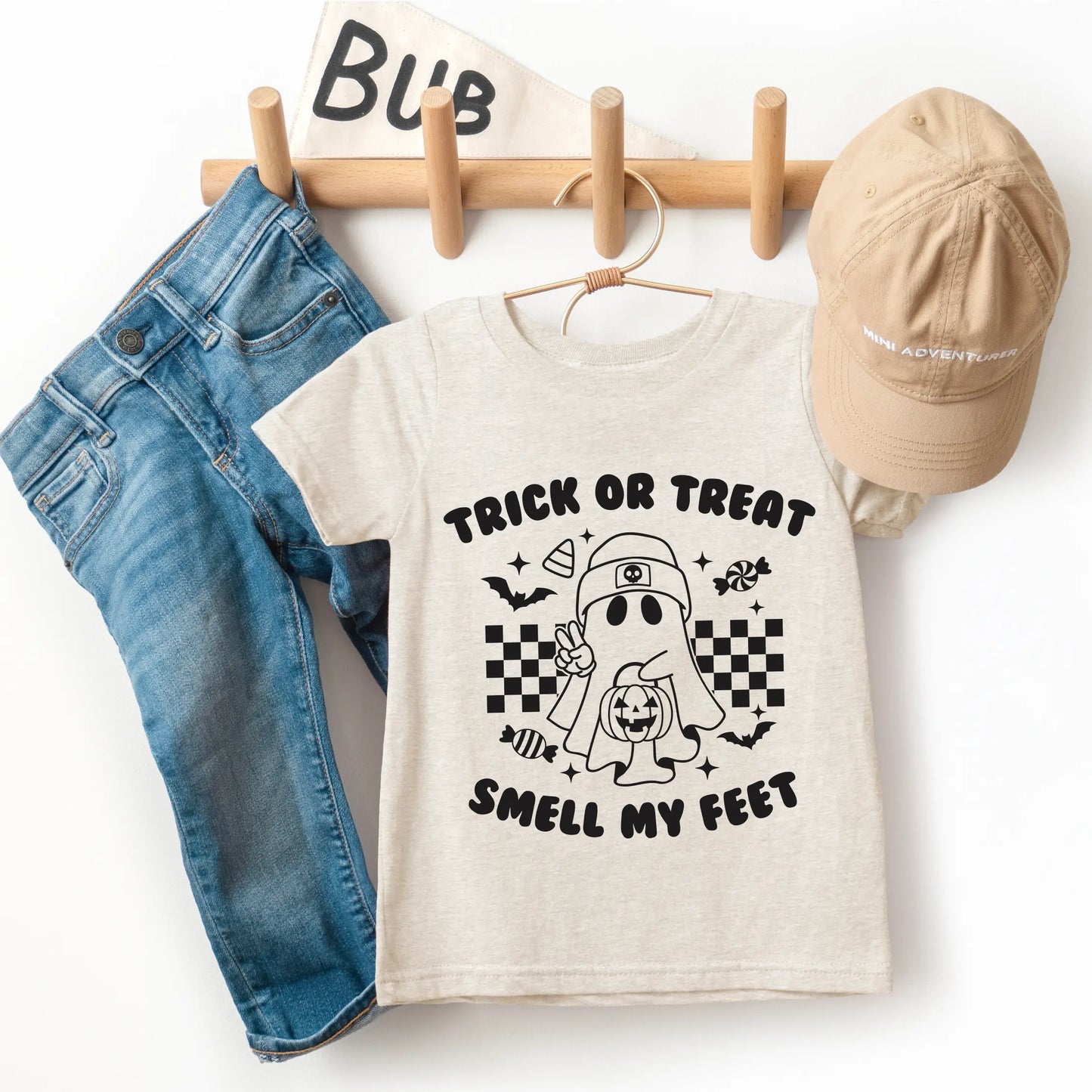 The Smell My Feet • Kids Tee is a beige unisex t-shirt featuring a cartoon ghost and Trick or Treat Smell My Feet text, made from super-soft cotton, shown with blue jeans and a beige cap on a wooden rack beside a “Bub” flag.