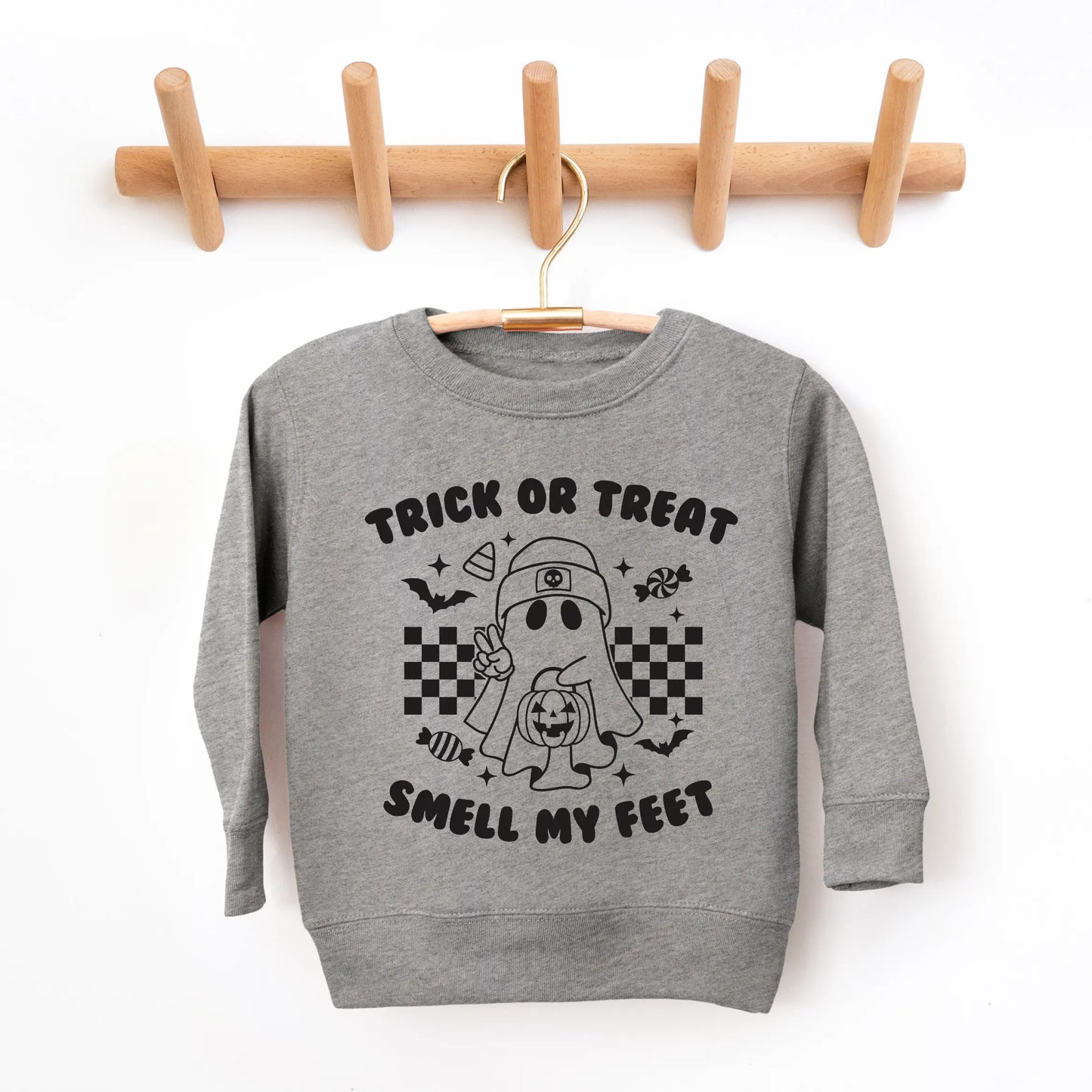 The Smell my Feet • Kids Pullover, a cozy gray unisex sweatshirt, hangs on a wooden rack. It features a cartoon ghost with candy, bats, stars, checkerboard patterns, and Trick or Treat Smell My Feet, perfect for Halloween-loving kids.