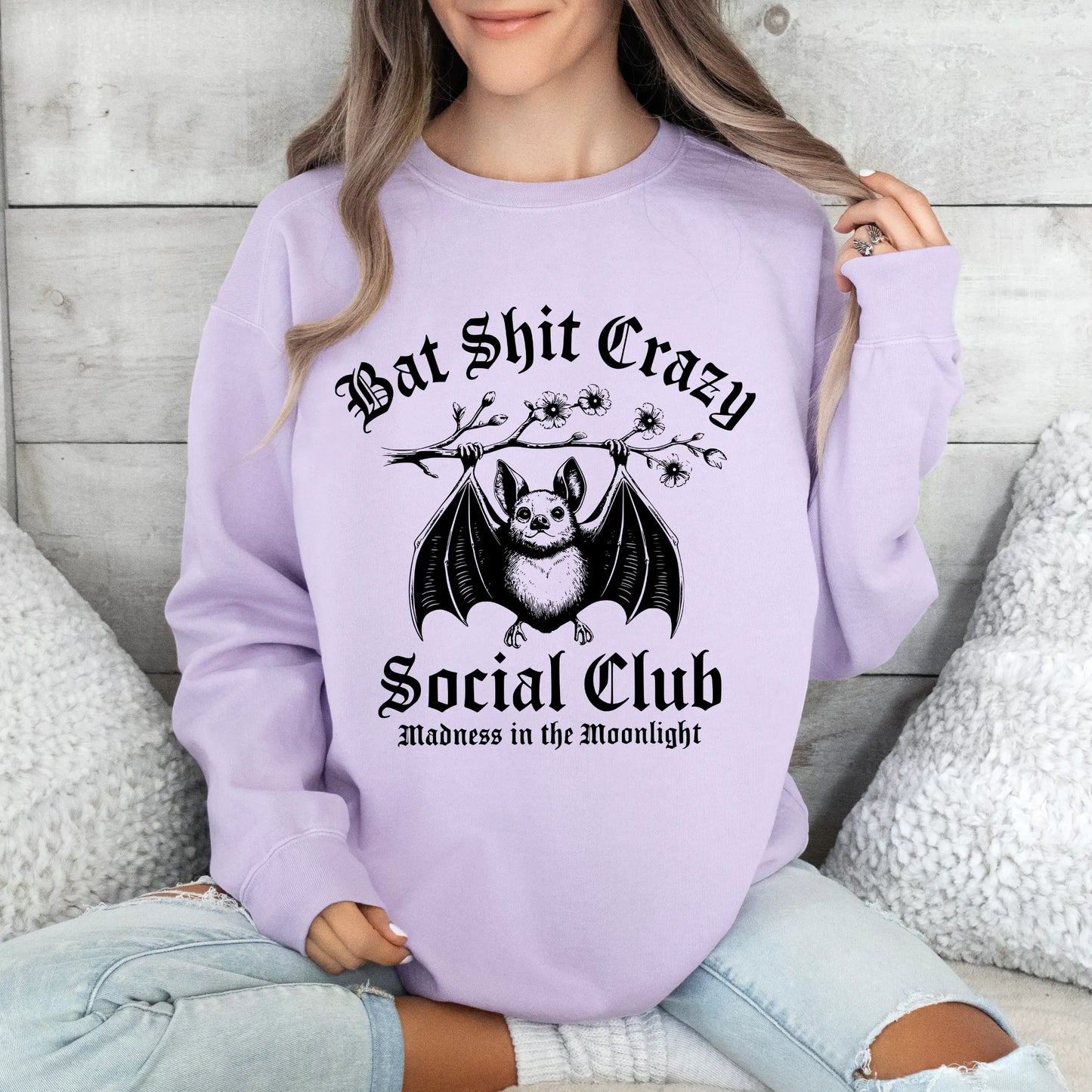 A woman sits cross-legged in a cozy gray setting, wearing the Crazy Social Club • Lilac Sweatshirt featuring a graphic bat and Bat Shit Crazy Social Club: Madness in the Moonlight text—an edgy, unisex cotton fleece style.