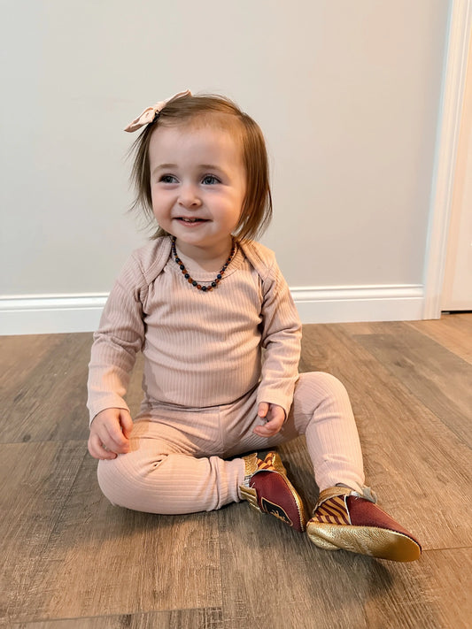 A toddler with light brown hair and a pink bow smiles while sitting on a wooden floor, wearing a light pink Ribbed Long Sleeve Bodysuit, a beaded necklace, maroon and gold shoes. A white wall and door are visible in the background.