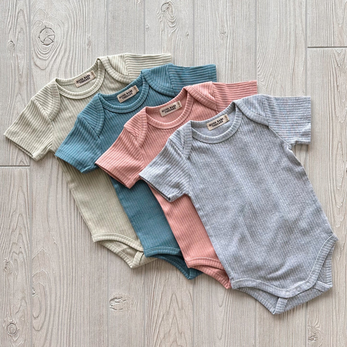 Four Ribbed Short Sleeve Bodysuits in beige, teal, pink, and light gray are displayed on a light wooden floor—an adorable addition to any baby clothing collection.