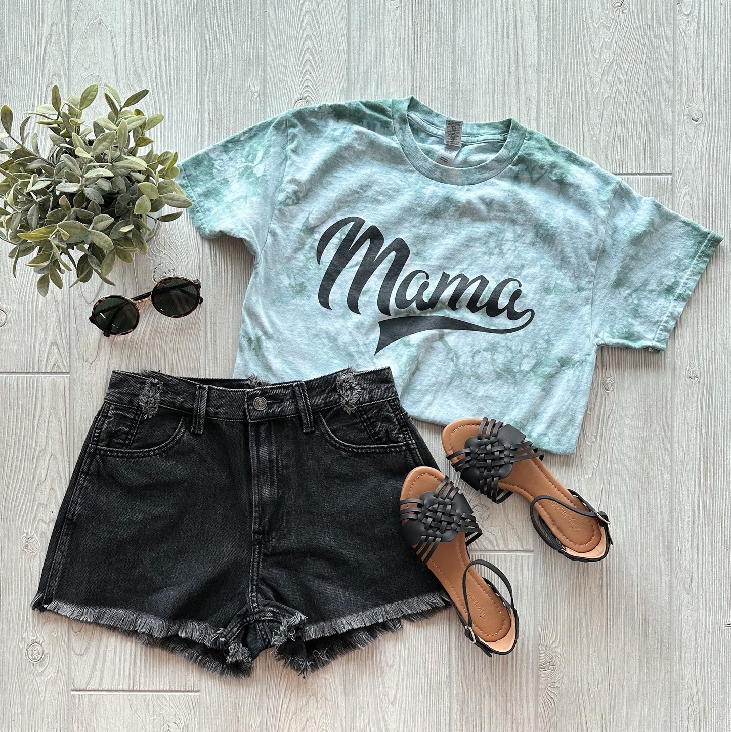 Flat lay of the Mama Sporty • Moss Tie Dye unisex t-shirt, paired with black frayed denim shorts, black woven sandals, round black sunglasses, and a small green potted plant on a light wood floor.
