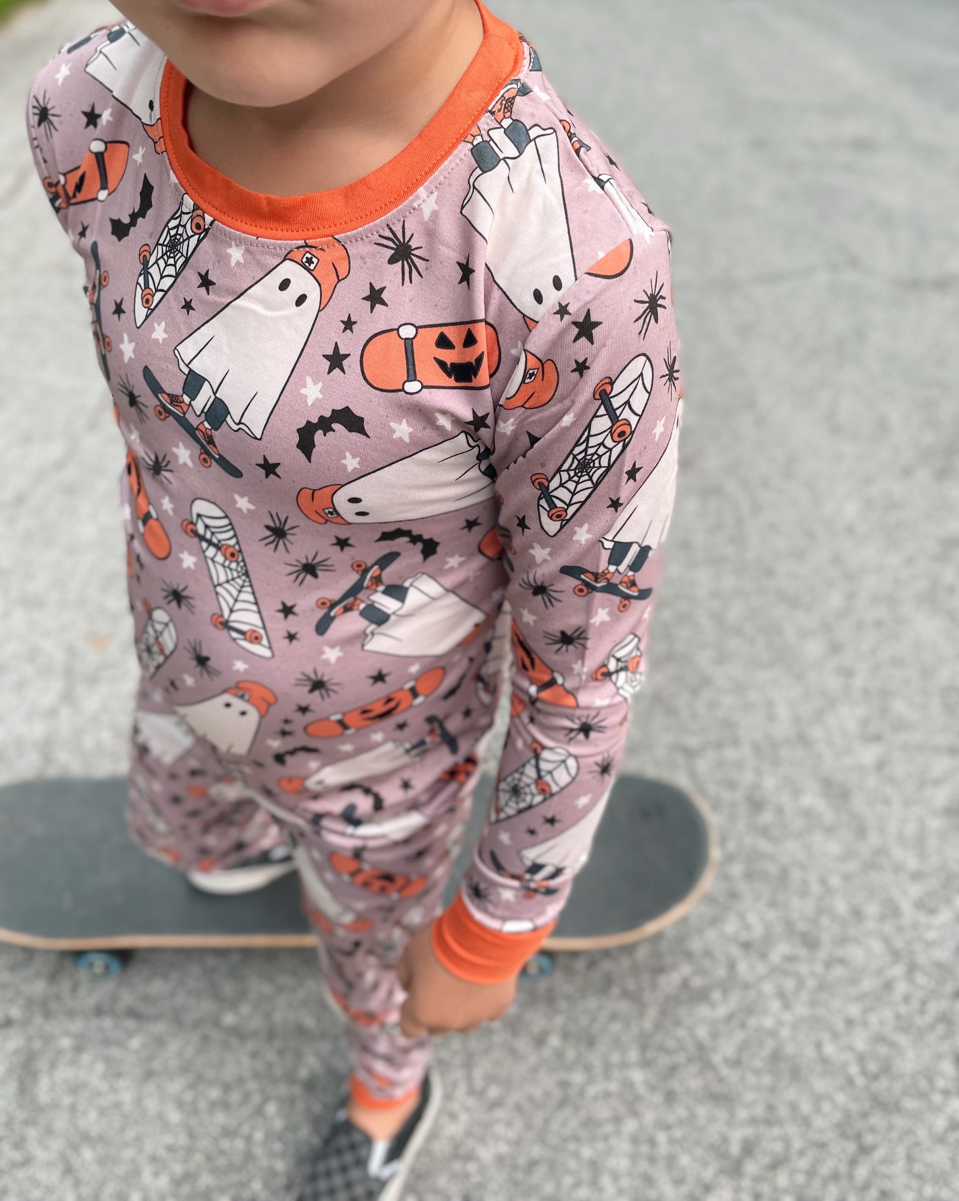 A child in our Skater Ghosts • 2 Piece Bamboo Set (Classic Fit)—Halloween pajamas with ghosts, pumpkins & bats—stands on a skateboard outside. The super-stretch bamboo fabric ensures cozy comfort; the childs face is partially out of frame.