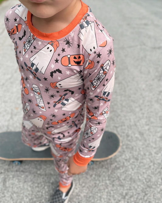 A child in our Skater Ghosts • 2 Piece Bamboo Set (Classic Fit)—Halloween pajamas with ghosts, pumpkins & bats—stands on a skateboard outside. The super-stretch bamboo fabric ensures cozy comfort; the childs face is partially out of frame.