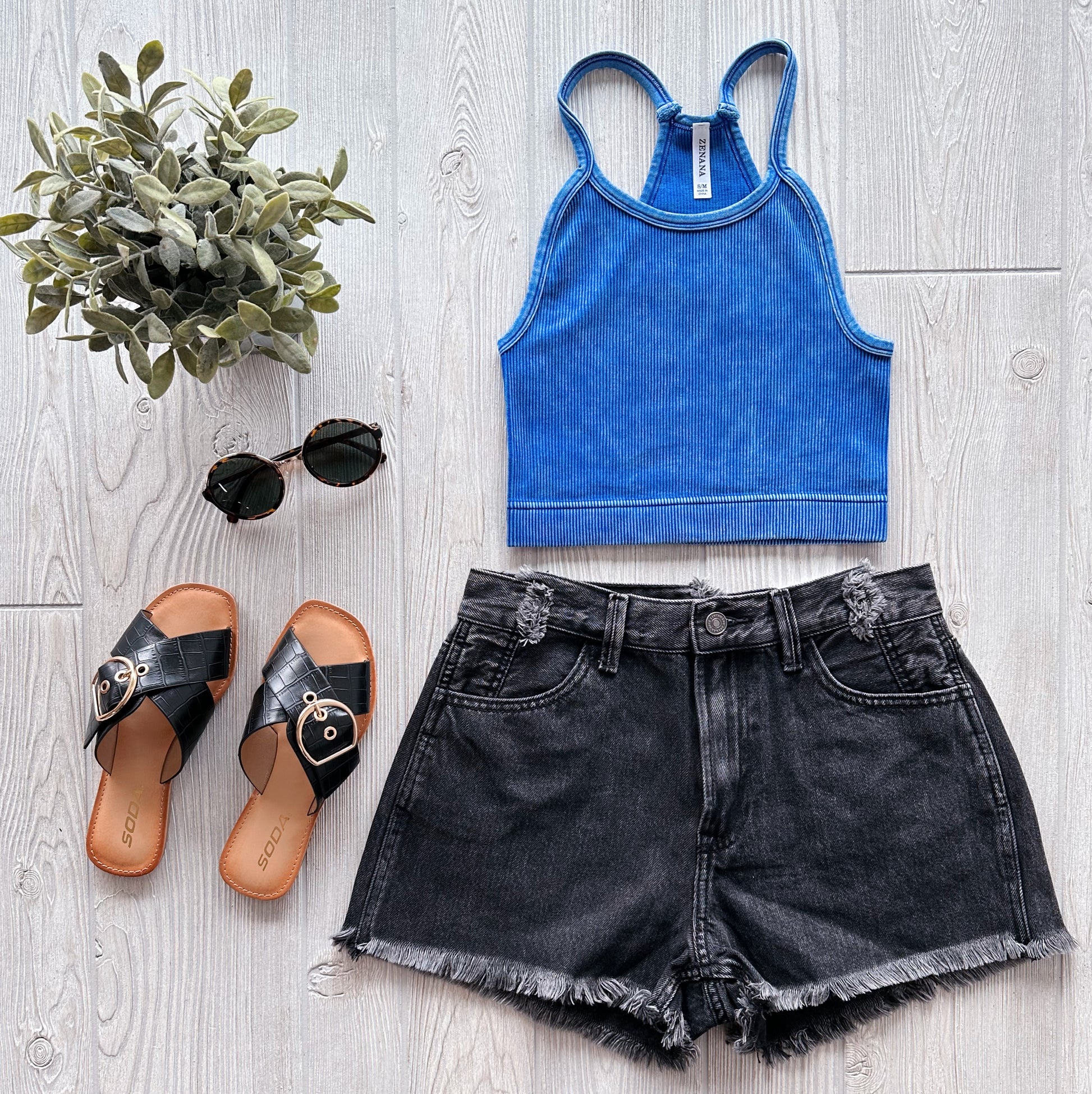 Flat lay of a casual summer outfit: a blue ribbed crop top, Lola Frayed Hem Shorts with an asymmetrical waistband, black slide sandals with gold buckles, round sunglasses, and a small potted plant on a light wood surface.
