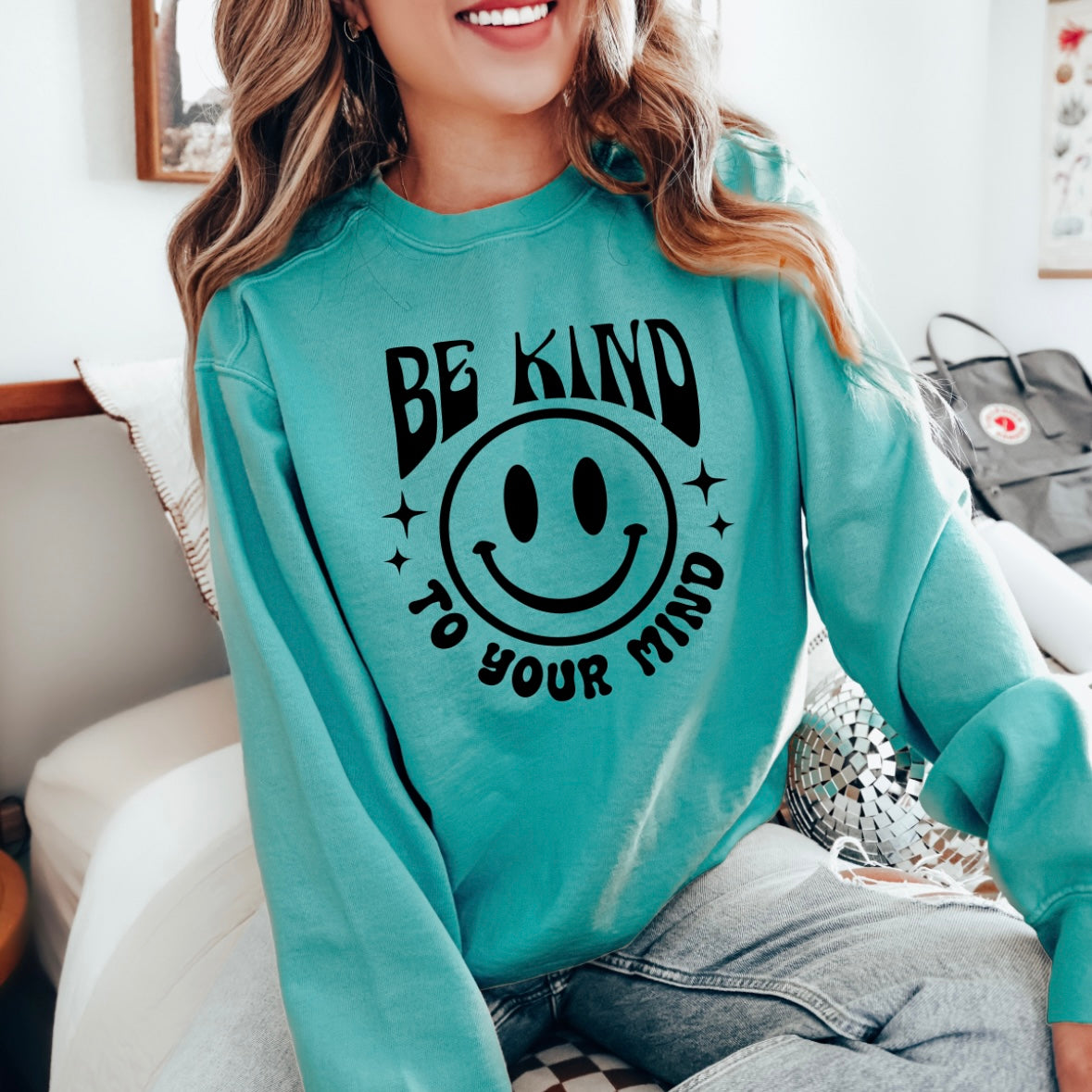 A cheerful person sits on a bed, smiling in a Seafoam Kind to your Mind long sleeve featuring a large smiley face graphic.