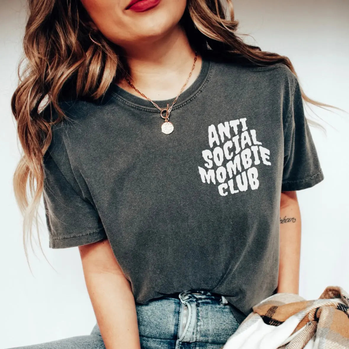 A woman wears the Anti Social Mombie • Comfort Tee, a faded black, garment-dyed T-shirt with white ANTI SOCIAL MOMBIE CLUB text. Shes styled with blue jeans and a necklace; only her torso and part of her face are visible.