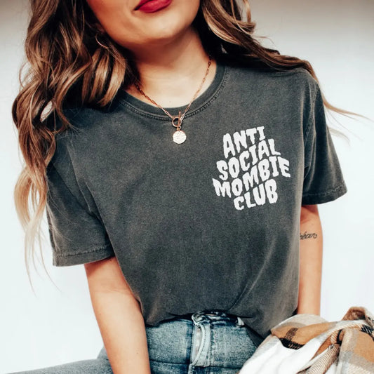 A woman wears the Anti Social Mombie • Comfort Tee, a faded black, garment-dyed T-shirt with white ANTI SOCIAL MOMBIE CLUB text. Shes styled with blue jeans and a necklace; only her torso and part of her face are visible.