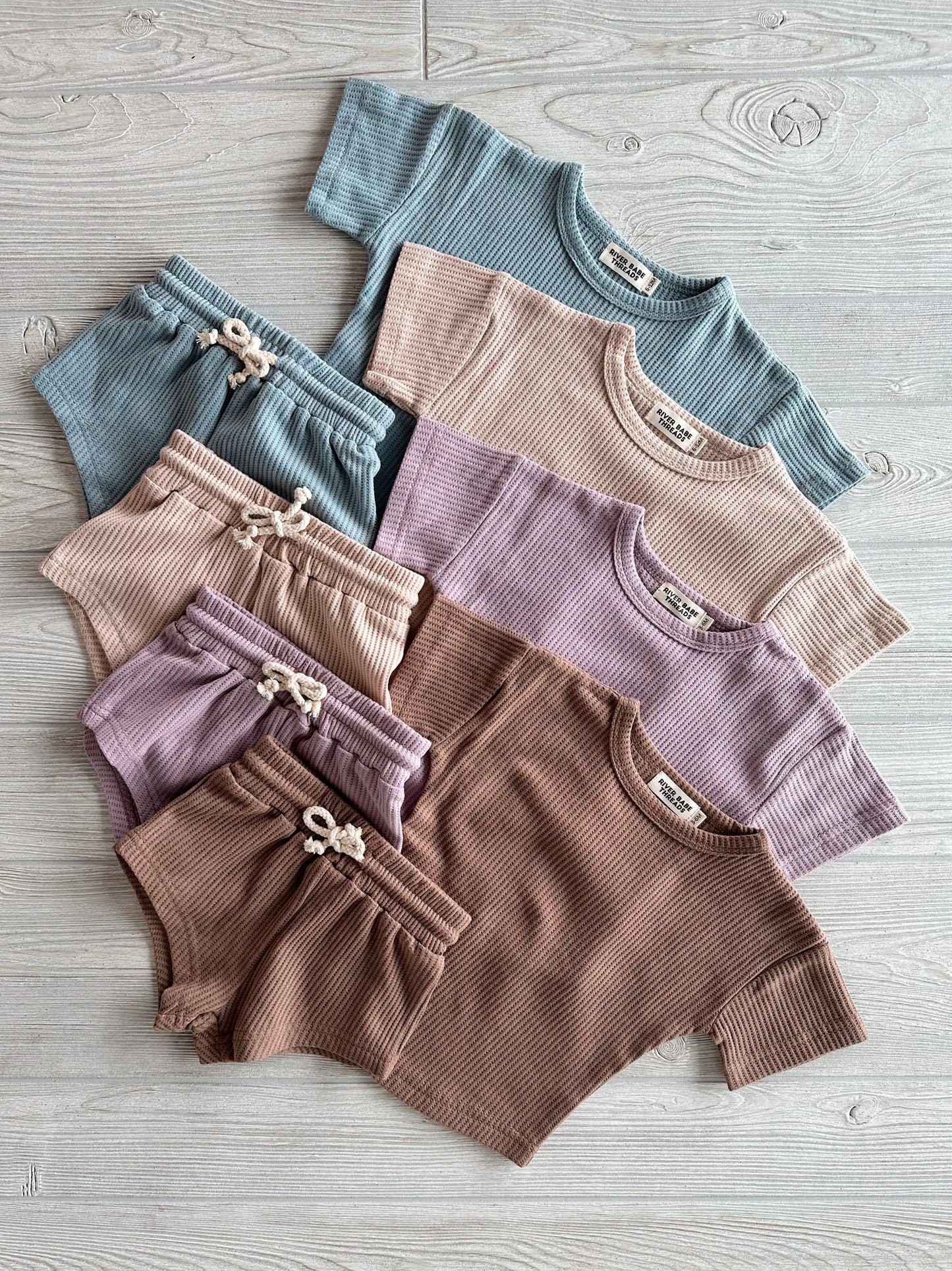A Steel Blue Waffle Shorty Set for babies and toddlers, featuring a ribbed organic T-shirt with matching shorts, is neatly displayed on a light wooden floor. This outfit fits true to size.