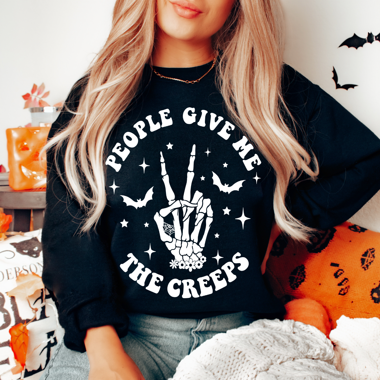 A woman with long, wavy blonde hair wears The Creeps • Black Pullover, a classic fit cotton/polyester sweatshirt featuring People give me the creeps with a skeleton hand, bats, and stars. Halloween decorations appear in the background.