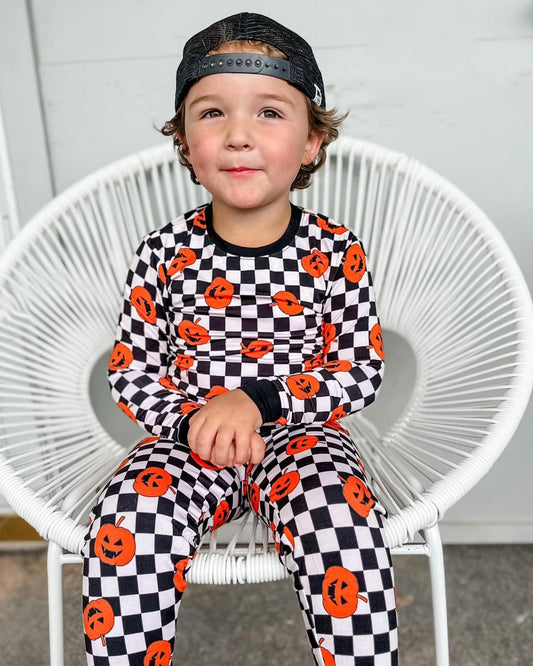 A young child sits on a white circular chair wearing a black baseball cap and the Checkered Pumpkins • 2 Piece Bamboo Set (Classic Fit), smiling slightly with hands resting on their lap.