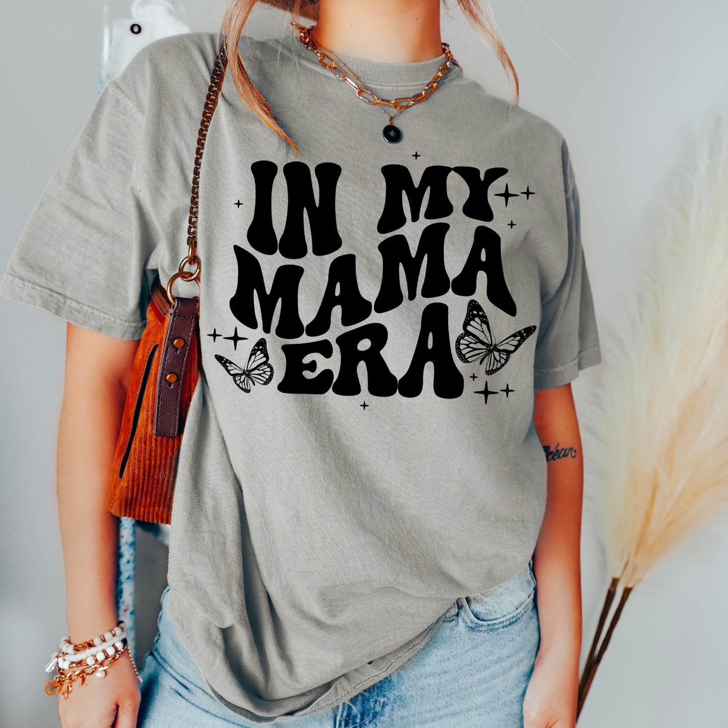 A person wears the Mama Era • Gray Dyed Tee featuring bold black IN MY MAMA ERA text with butterfly and sparkle graphics. This ringspun cotton unisex shirt offers a comfortable fit, styled with a brown purse and jewelry.
