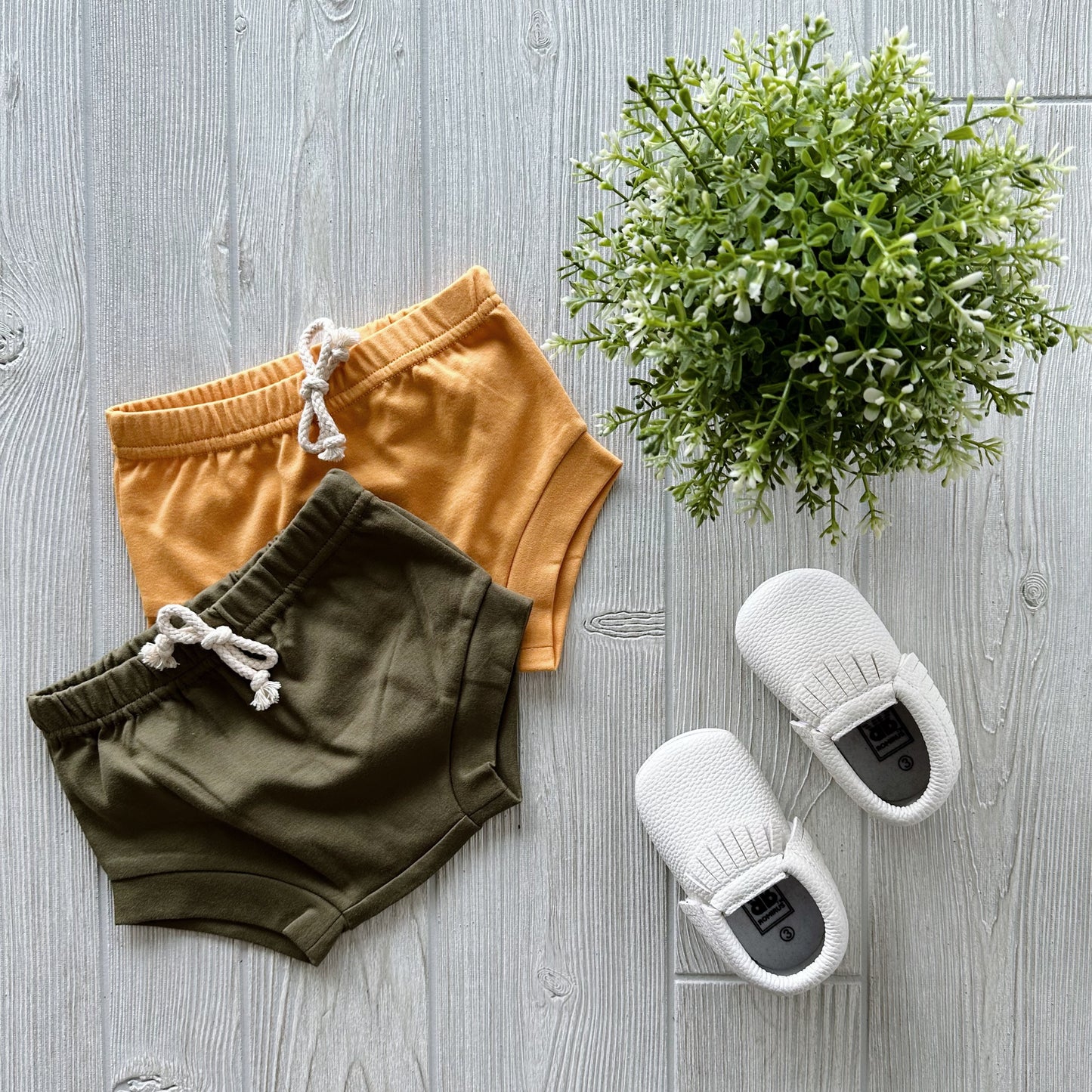 Flat lay on light wood: Organic Cotton Bummies in mustard yellow, olive green shorts with faux drawstring, white knit baby shoes, and a small green potted plant.