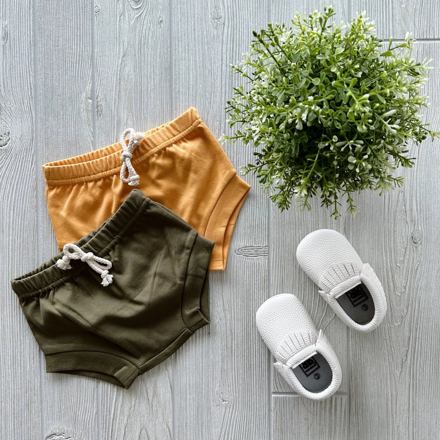 Flat lay on light wood: Organic Cotton Bummies in mustard yellow, olive green shorts with faux drawstring, white knit baby shoes, and a small green potted plant.