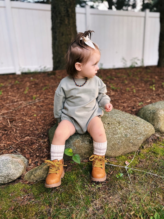 A toddler with a light bow sits on a large rock outdoors, wearing the French Terry Bubble Romper, tall cream socks, and brown boots. They look to the side with a white fence and trees in the background.