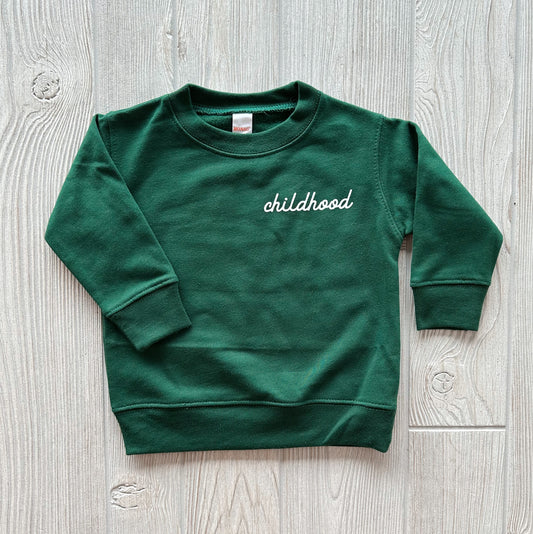 The Childhood • Forest Infant Pullover is a dark green, oversized long-sleeve sweatshirt with childhood in white cursive on the left chest, shown laid flat on a light wood surface.