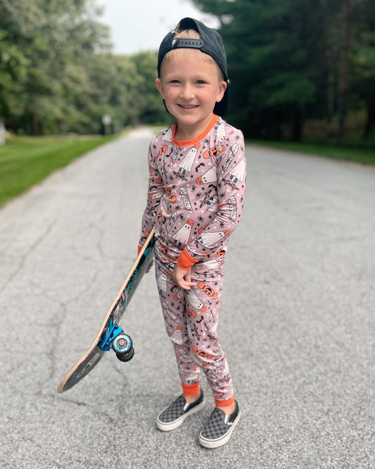 A young boy in the Skater Ghosts • 2 Piece Bamboo Set (Classic Fit) and a backward cap smiles on a quiet road with a skateboard, surrounded by trees and grass.
