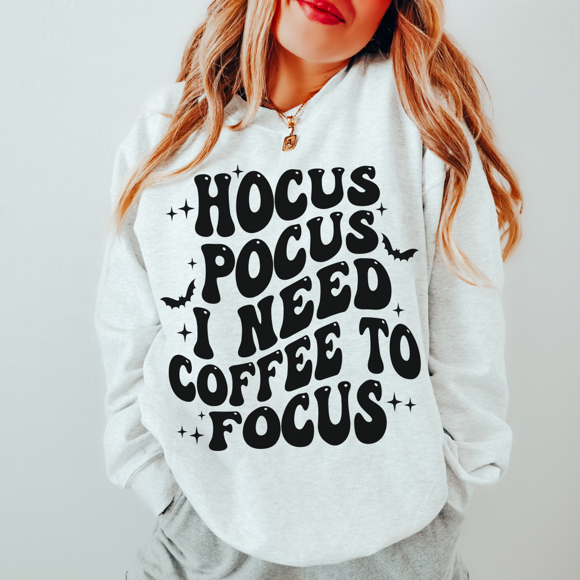 Woman with long hair wearing the Coffee to Focus • Ash Pullover, a cotton/polyester sweatshirt featuring “HOCUS POCUS I NEED COFFEE TO FOCUS” in bold black letters with bat and sparkle graphics around the text.