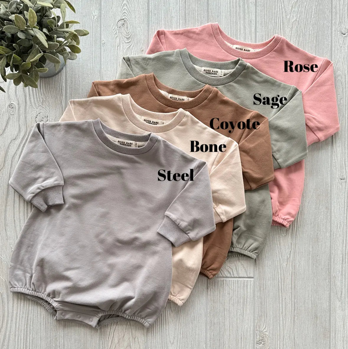 Five French Terry Bubble Rompers in muted shades (Steel, Bone, Coyote, Sage, Rose) are arranged overlapping on a light wooden surface with a small green plant. Each romper is labeled by color—a perfect cool weather staple.