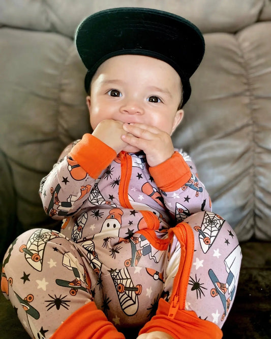 A baby with sensitive skin wears the Skater Ghosts • Bamboo Zippy (Classic Fit), smiling on a couch with a black baseball cap worn backward and holding their hands to their mouth.